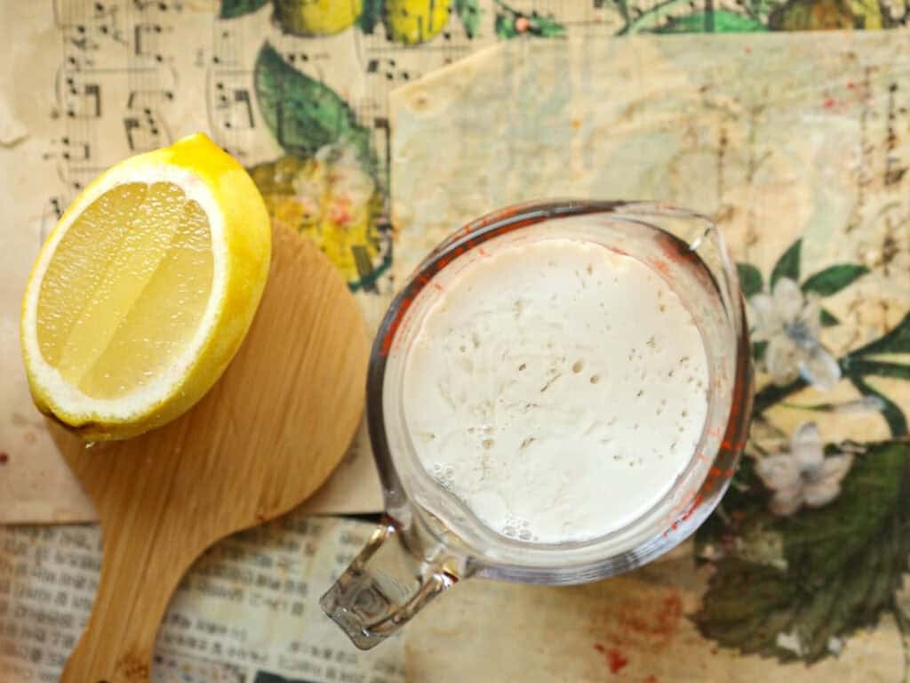 A sliced lemon on a wooden board next to a glass measuring cup filled with a frothy liquid. The background features a botanical pattern with flowers and out-of-focus sheet music.